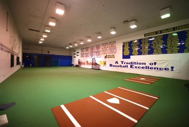 An indoor baseball training facility with green turf, multiple pitching mounds, and achievement banners on the walls.