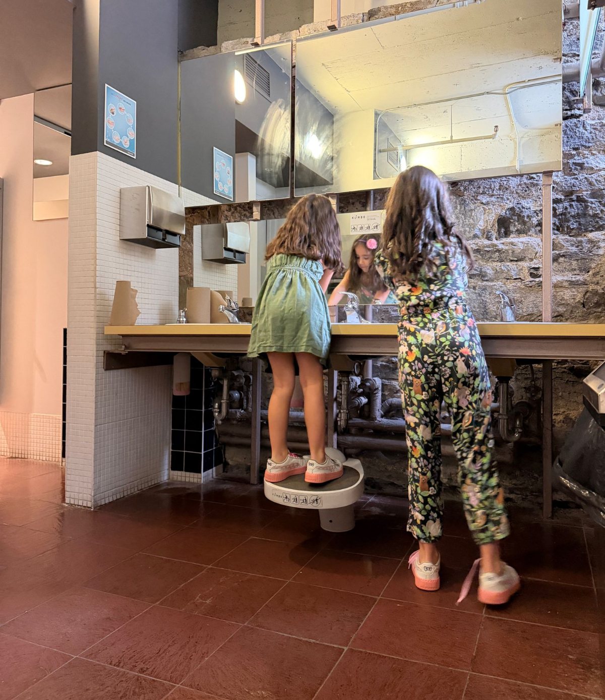 Two girls at a public restroom counter, one standing on a Klimbo K01
