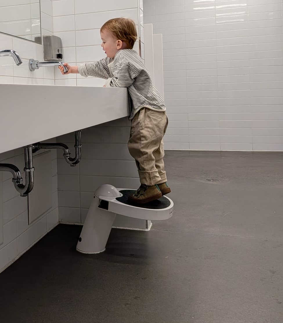 A toddler standing on a Klimbo step stool reaching the faucet of an adult-height sink