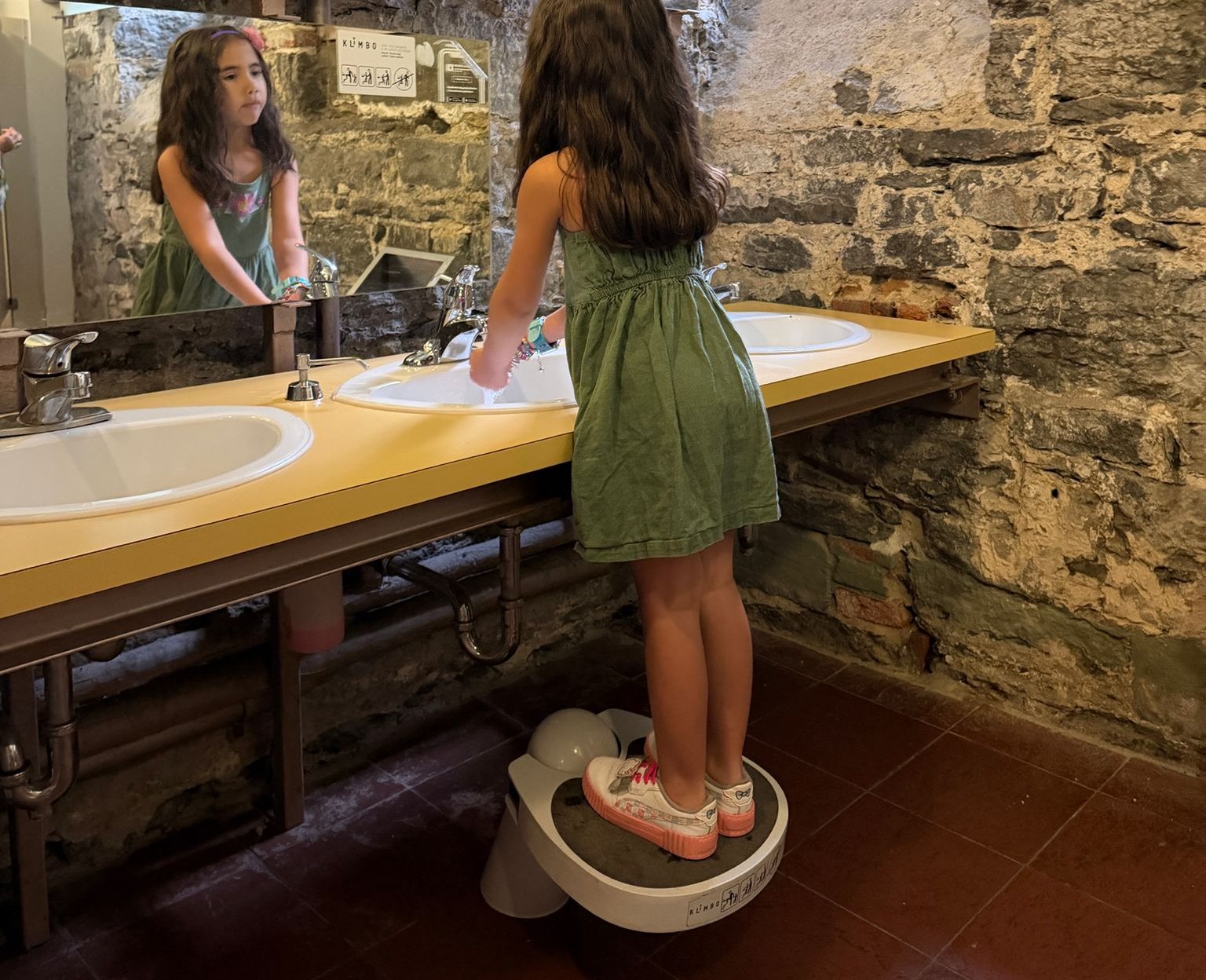 A girl standing on a Klimbo K01 step stool washing her hands at a public restroom sink