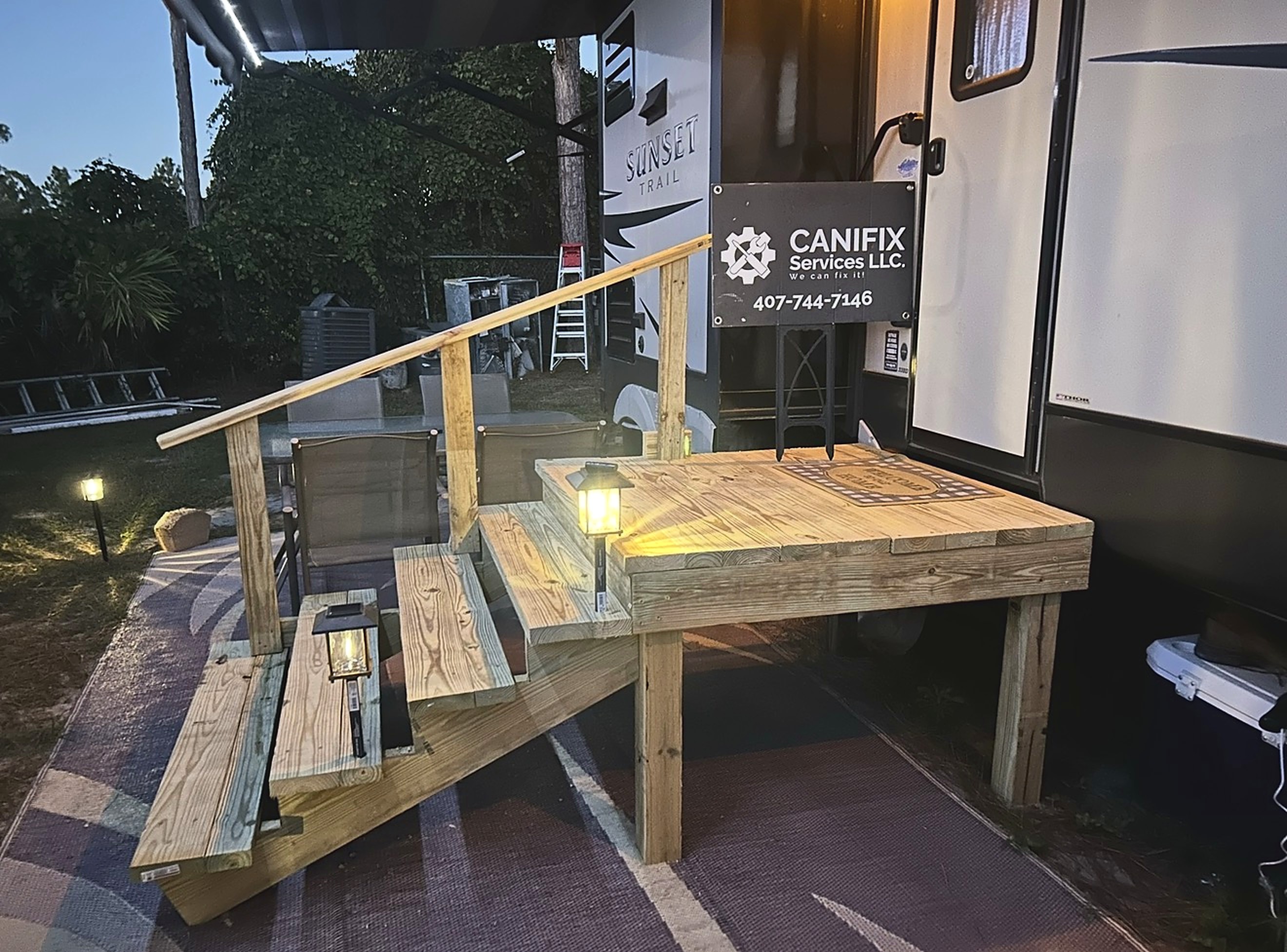 New pressure-treated wood landing with stairs and a handrail at an RV door at dusk, lit by small lanterns, with a CANIFIX Services LLC yard sign showing 407-744-7146.