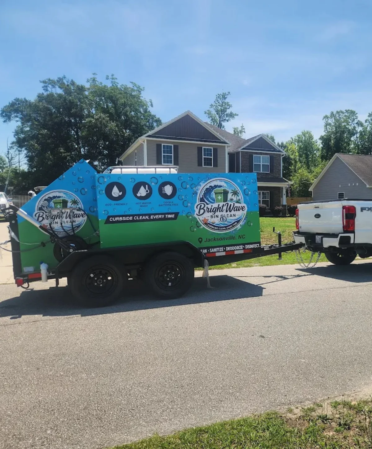 A Bright Wave Bin Clean service trailer, green and blue, hitched to a white pickup truck on a residential street.