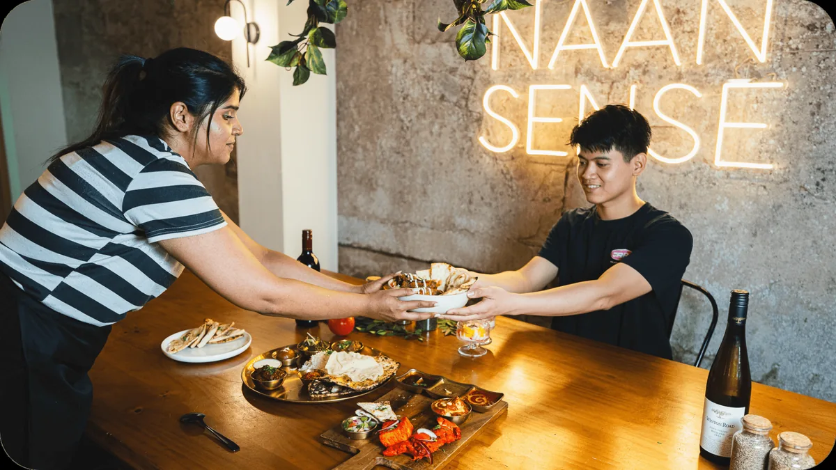 Staff handing a rice bowl to a smiling guest, thali on the table, under the Naan Sense neon sign, Newtown