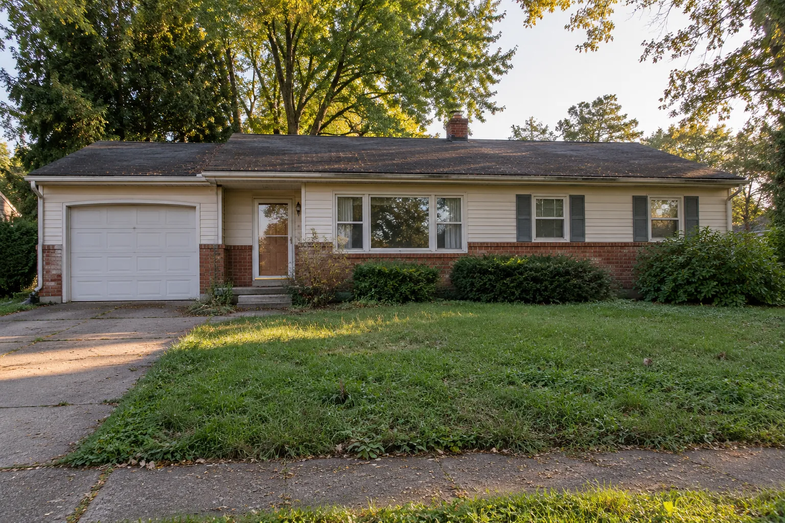 A single-story 1970s ranch house with an attached garage, an older roof and an overgrown lawn