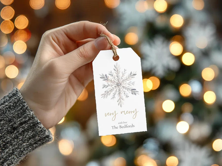 A hand holding a watercolor snowflake gift tag that reads very merry, with love, The Bedfords