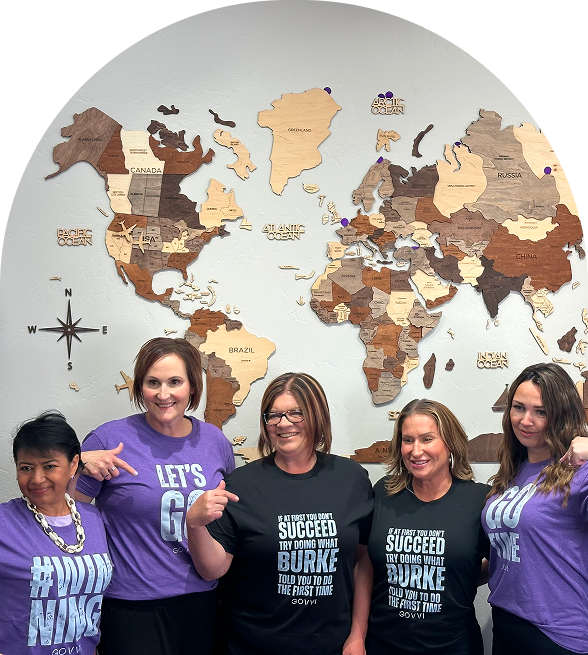 Five women standing in front of a decorative wooden world map, smiling and pointing at their motivational team shirts, representing unity, leadership, and empowerment in the Go Time Social community