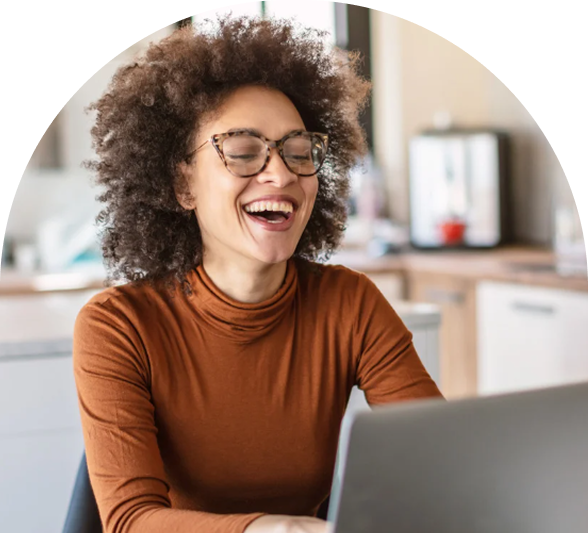 Woman with curly hair and glasses smiling while working on a laptop in a kitchen setting