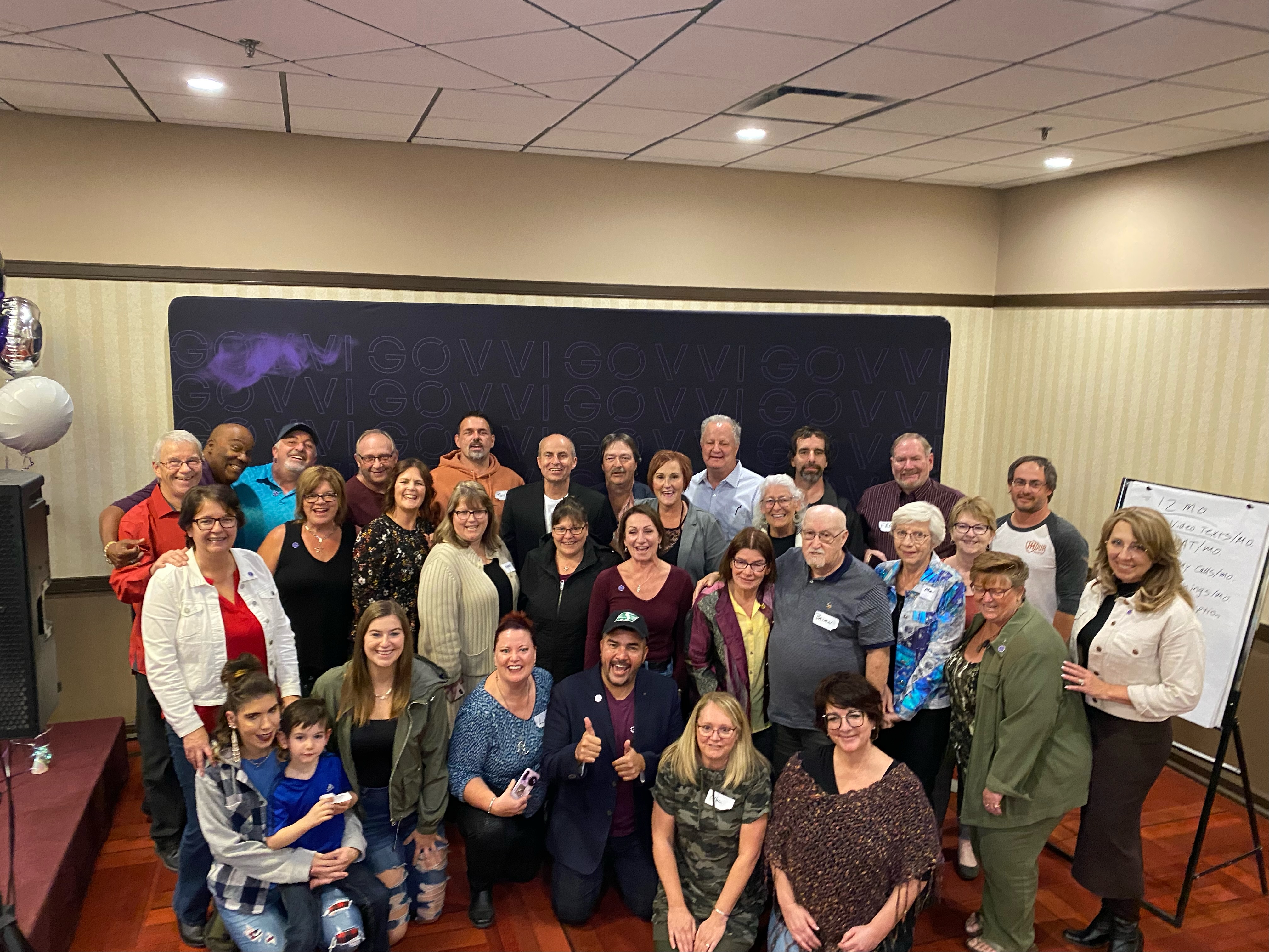 Group photo of dozens of people smiling in a conference room with a presentation board behind them