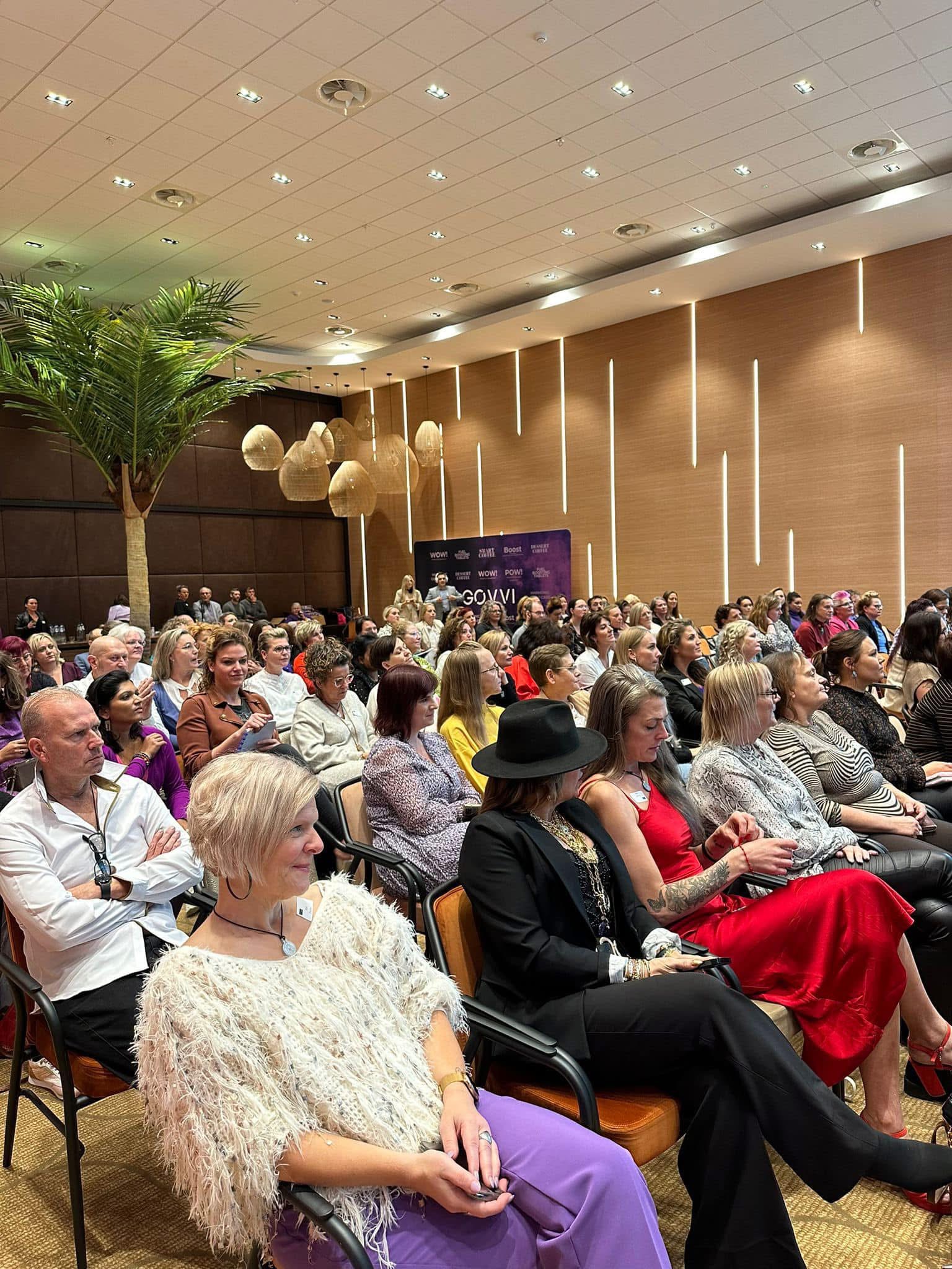 Large seated audience attending an event in a room decorated with balloons and plants