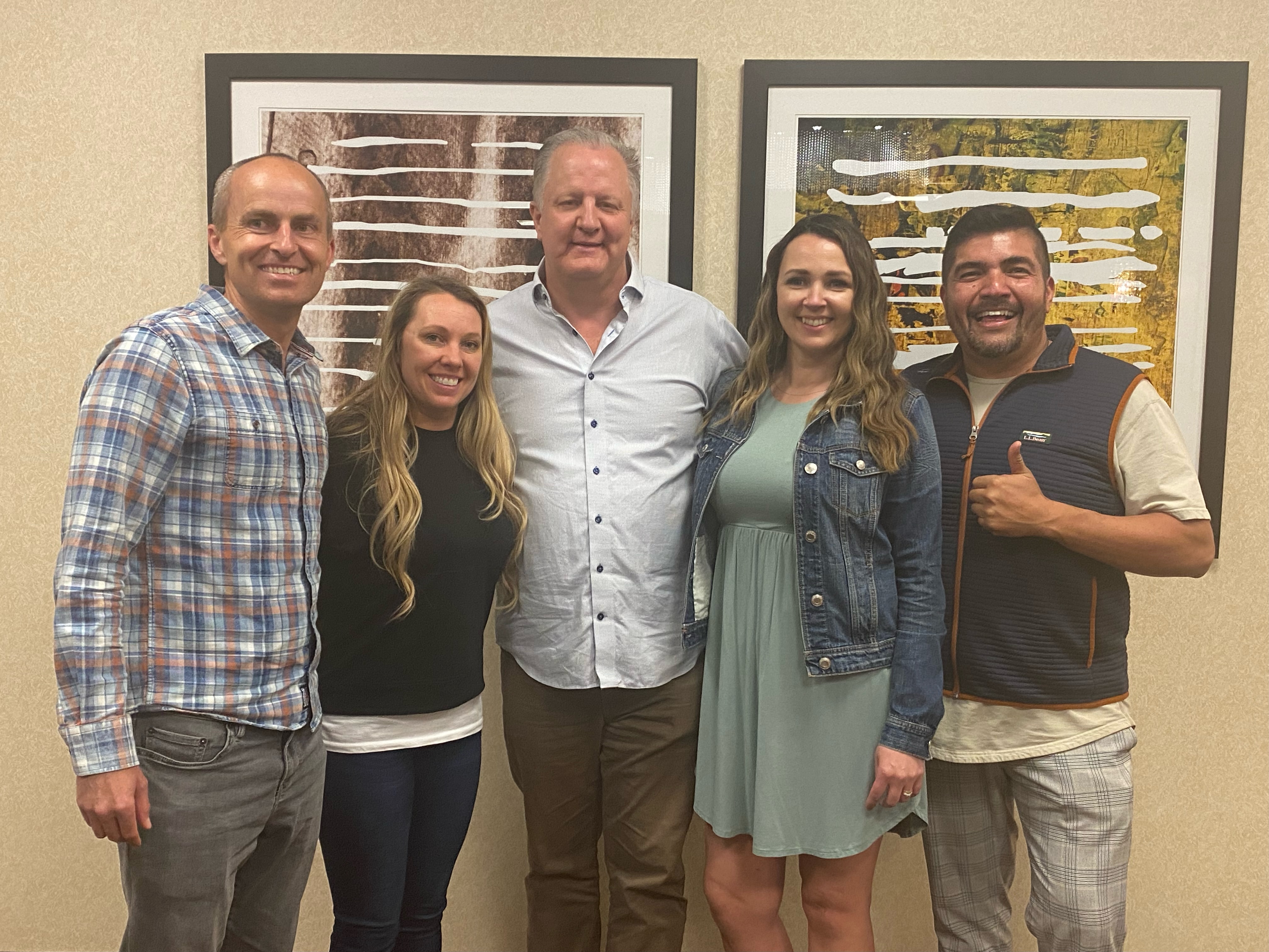 Group of five adults smiling and posing indoors in front of framed art