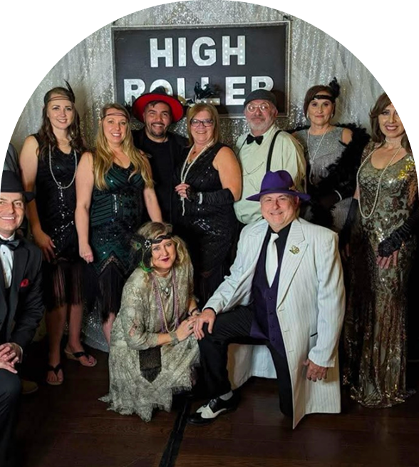 Group of people dressed in 1920s-themed attire posing in front of a “High Roller” sign at a costume party or themed event
