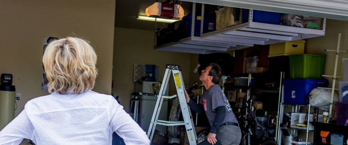 Garage door technician servicing an overhead opener while the homeowner watches from inside the garage.