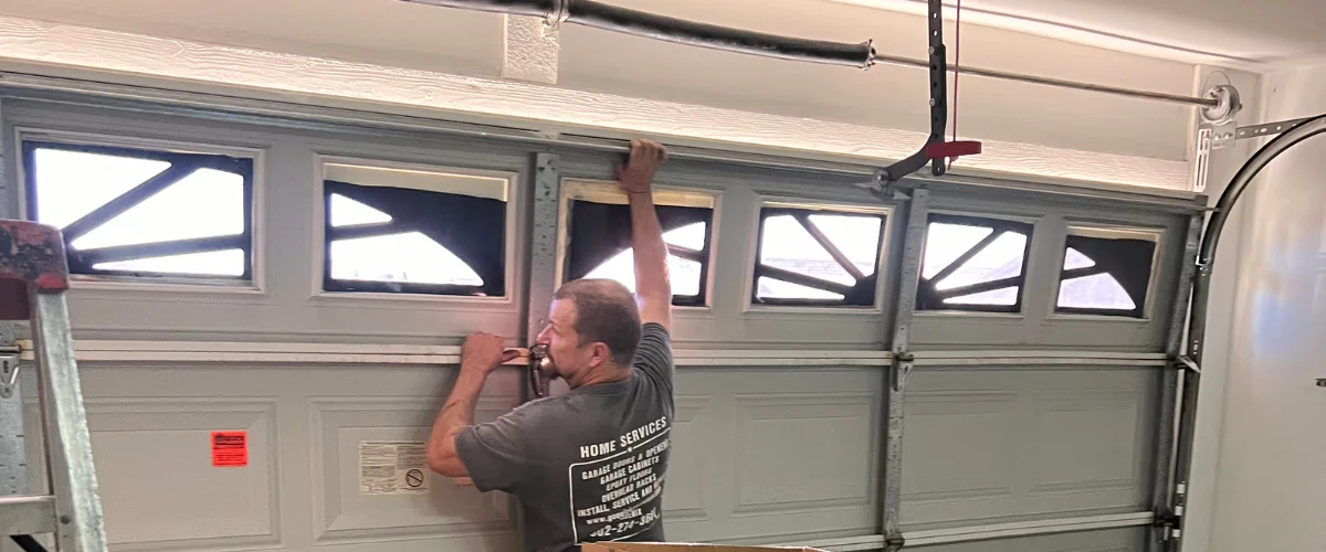 A man wearing a grey Home Services t-shirt installing decorative window inserts into a light grey garage door.