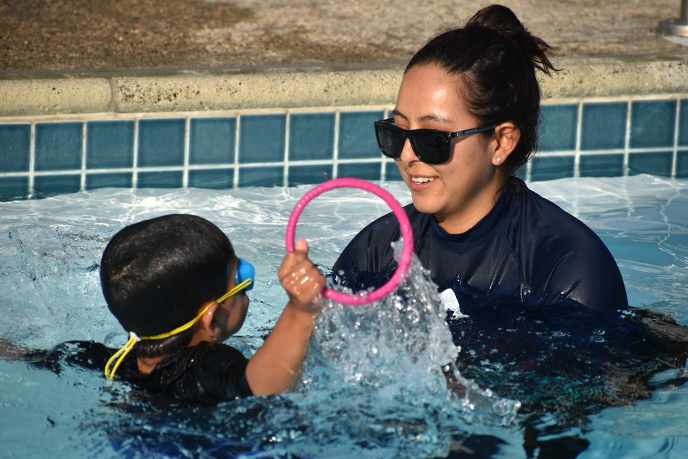 Child with pink ring enthusiastically engaging in water with instructor