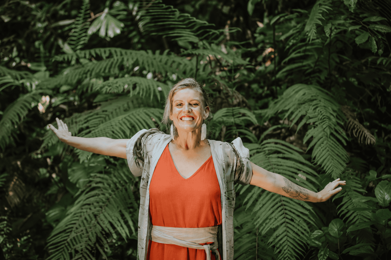 Olivka standing with her arms wide open in front of dense green ferns, wearing a poppy-orange dress.