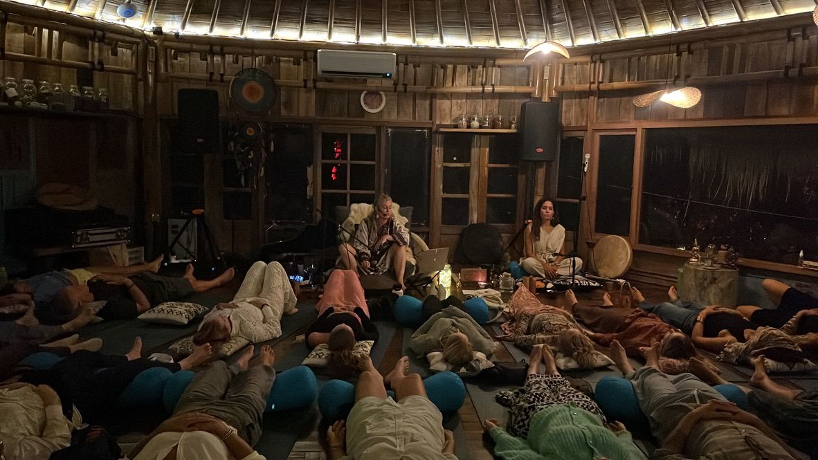 A round bamboo hall at night, a circle of people lying down with their eyes closed while Olivka and a second musician play.
