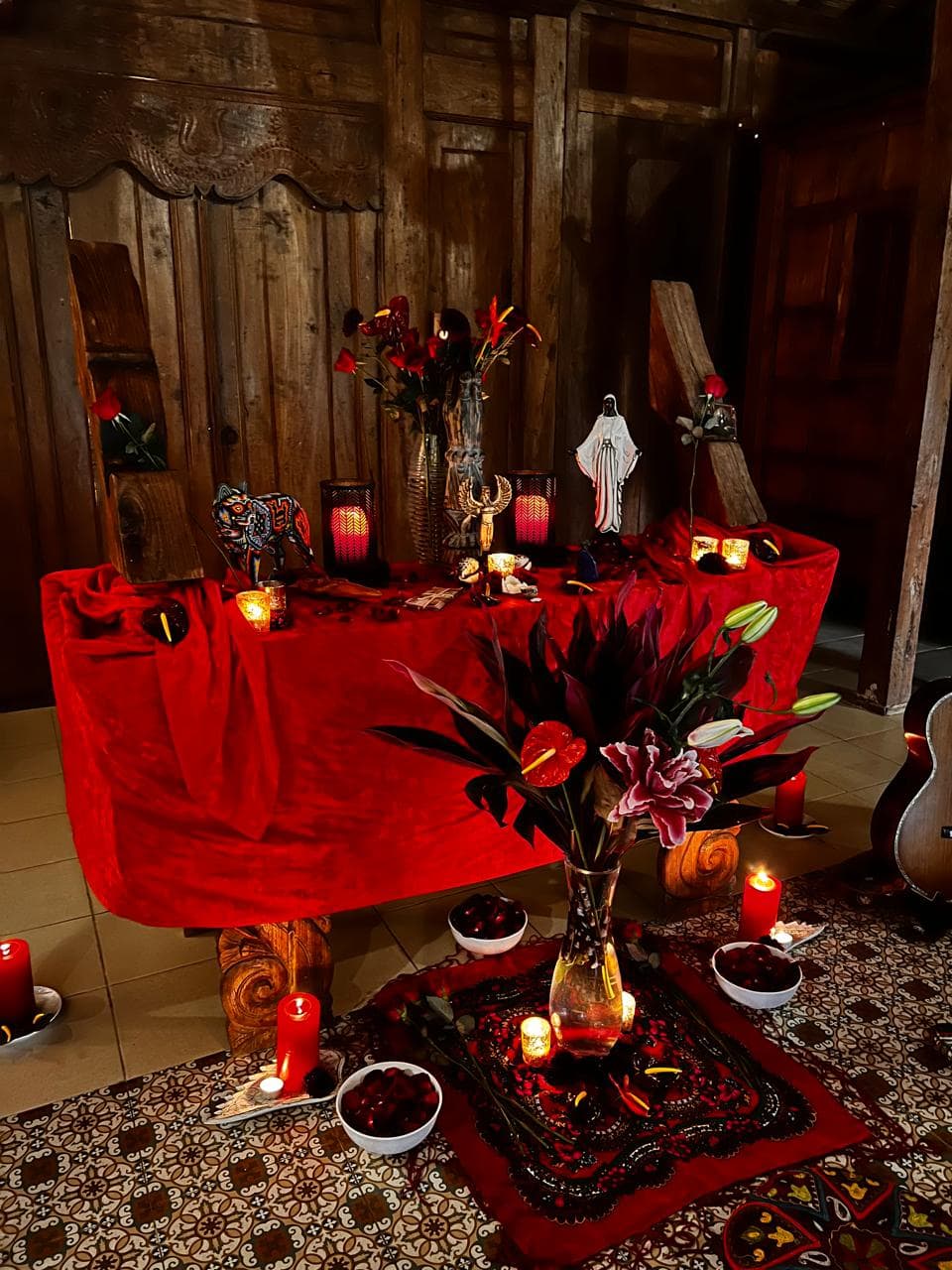 A red velvet altar by candlelight, set with roses, anthuriums, lilies and bowls of fruit.