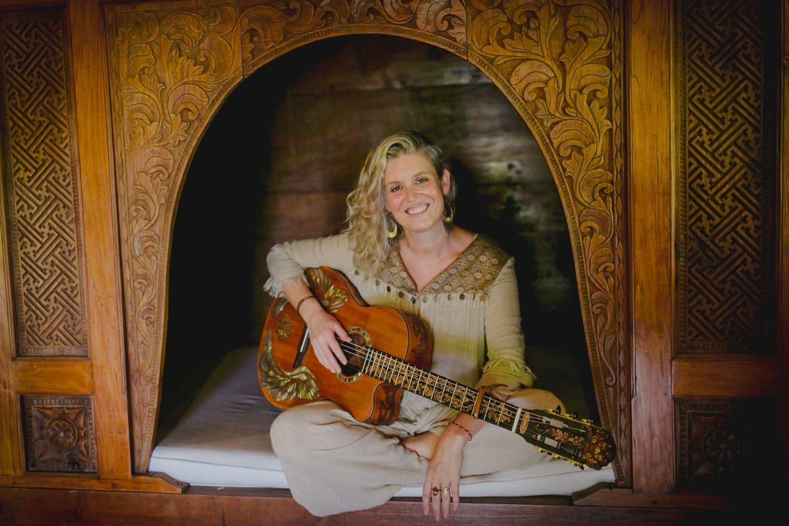 Olivka sitting cross-legged in a carved wooden alcove, smiling, holding an inlaid guitar.