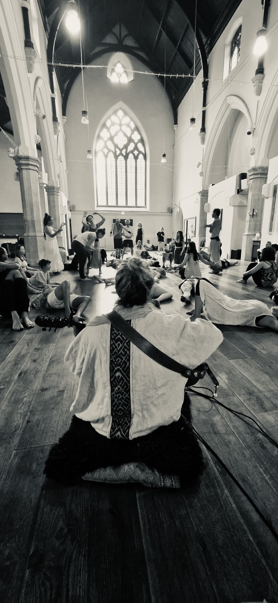 Olivka seated on the floor of a church hall with her guitar, a room of people moving and resting around her.