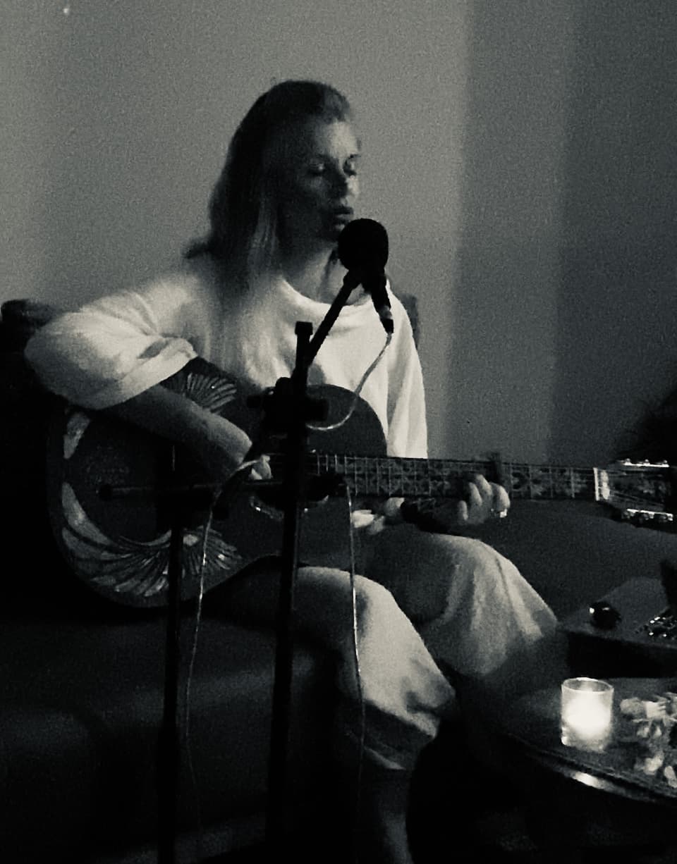 Olivka singing into a microphone on a sofa by candlelight, her guitar across her lap.