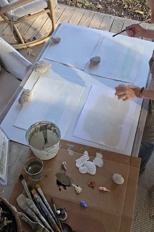 "Overhead view of an artist's hands painting soft abstract washes on paper weighted down with stones, in the outdoor studio of Dallas-based artist Sonya Edwards."