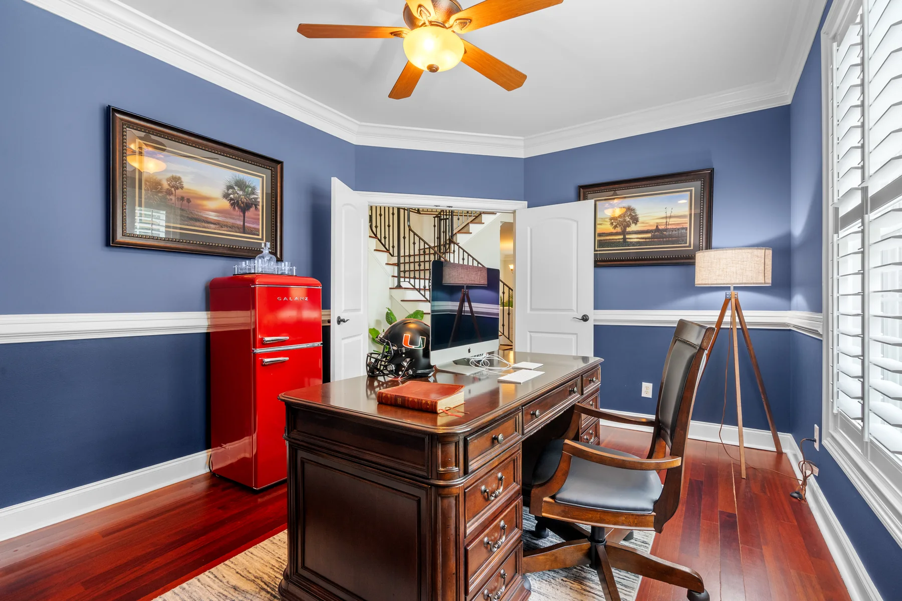 Room with blue walls, a wooden desk, framed pictures, and a ceiling fan