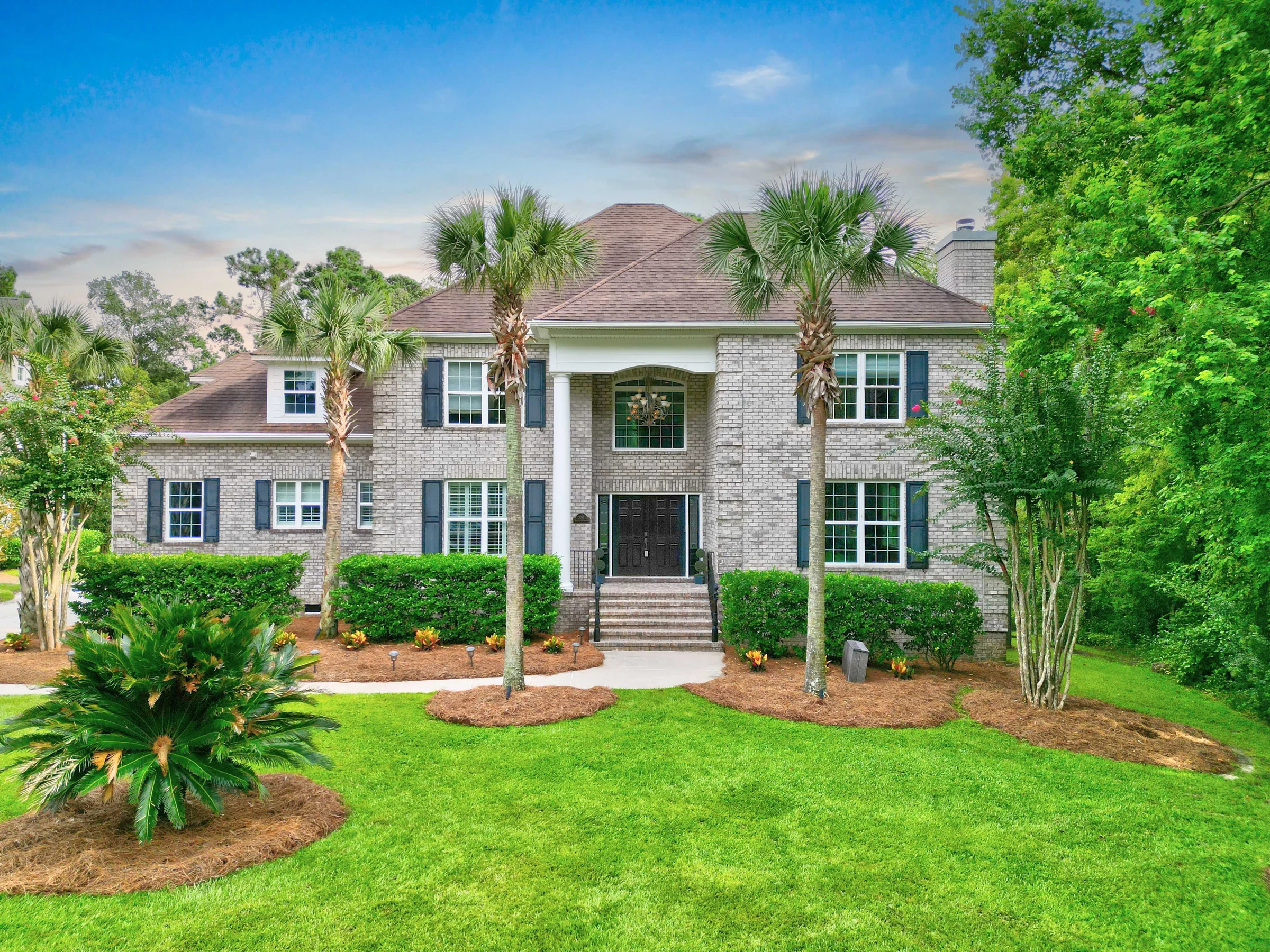 Large brick home with a brown roof, blue shutters, and palm trees