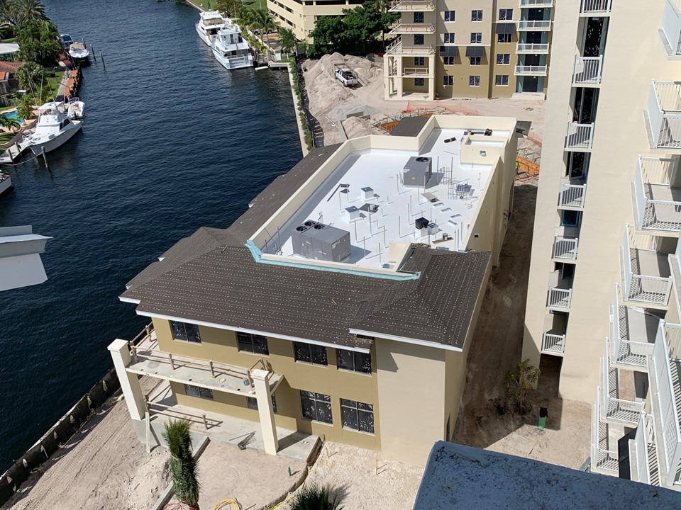 Aerial view of a building where a sloped shingle roof section meets a flat roof section, with a high-rise tower beyond