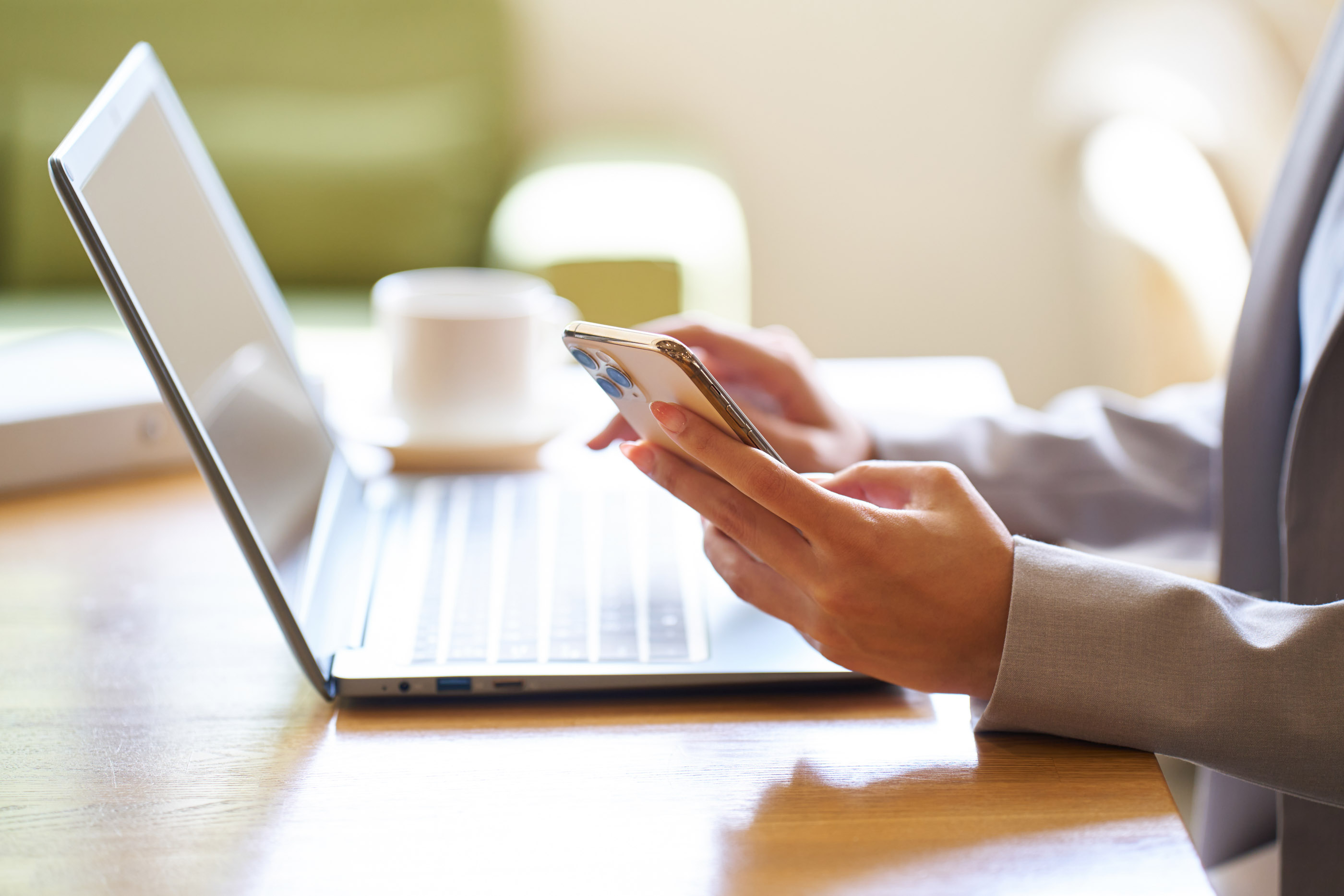 Business owner checking a smartphone beside an open laptop and a coffee cup on a wooden desk