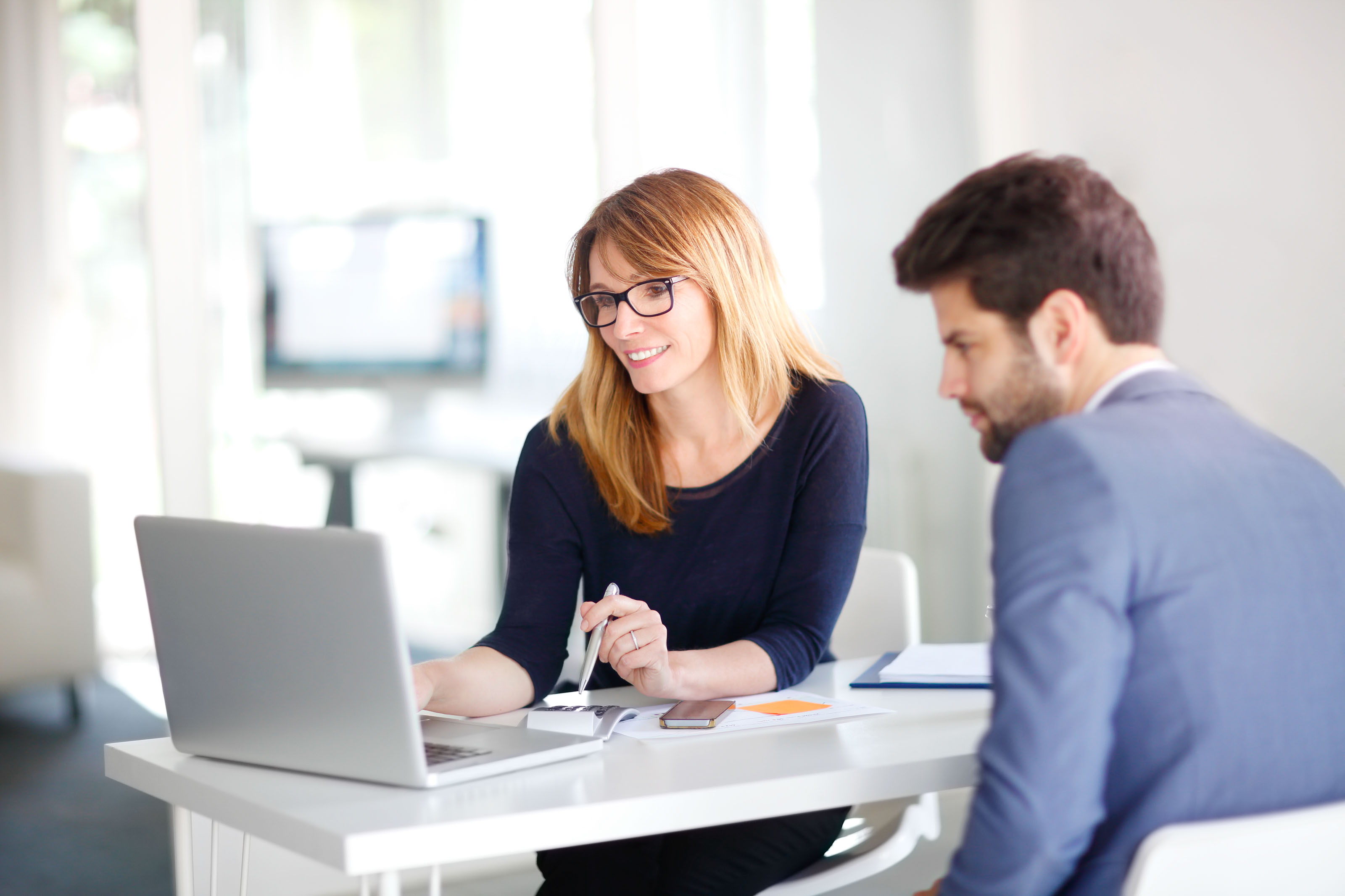 A woman in glasses reviews notes on a laptop with a man in a blue suit at a bright white office desk