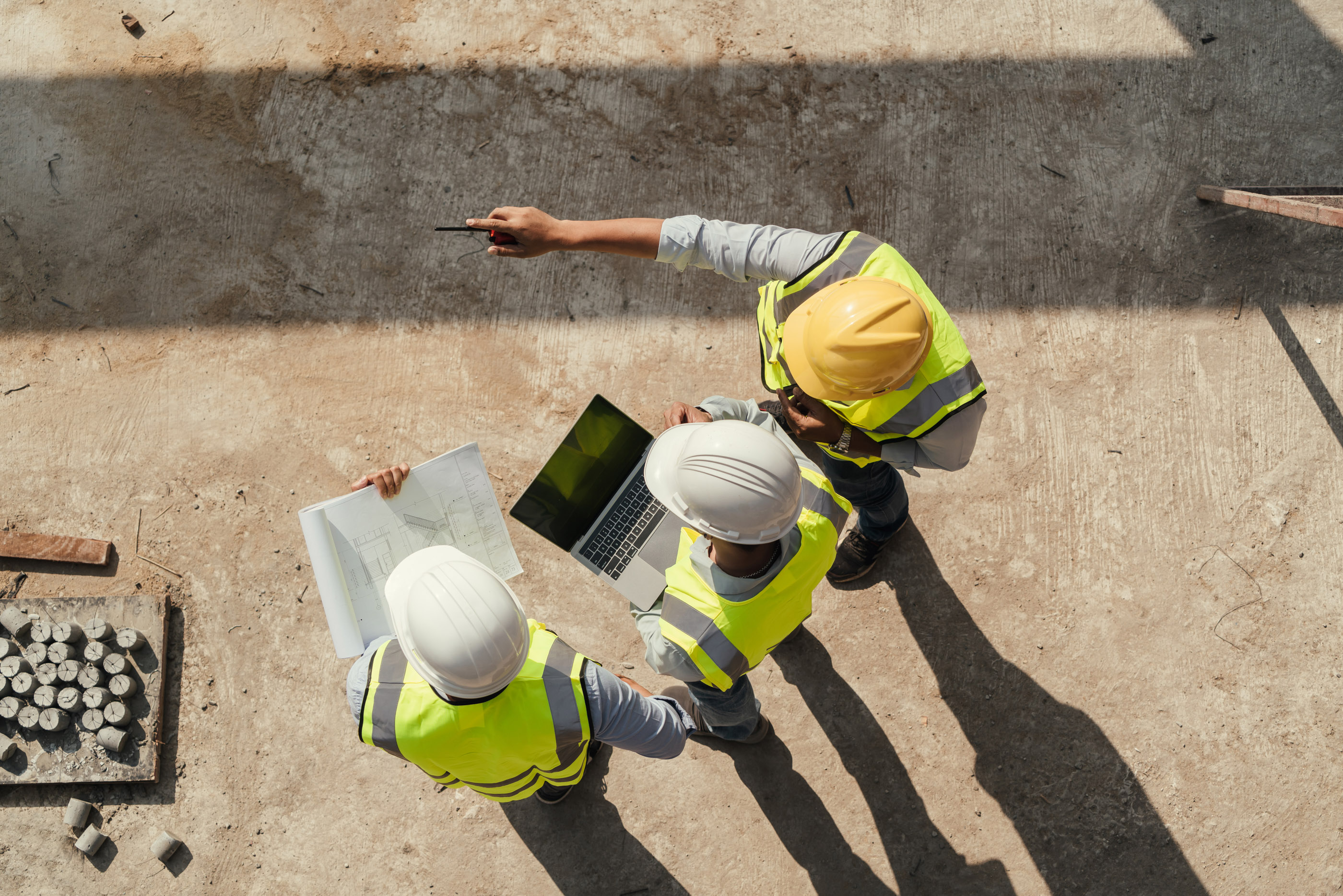Three construction workers in high-visibility vests reviewing blueprints and a laptop, seen from above