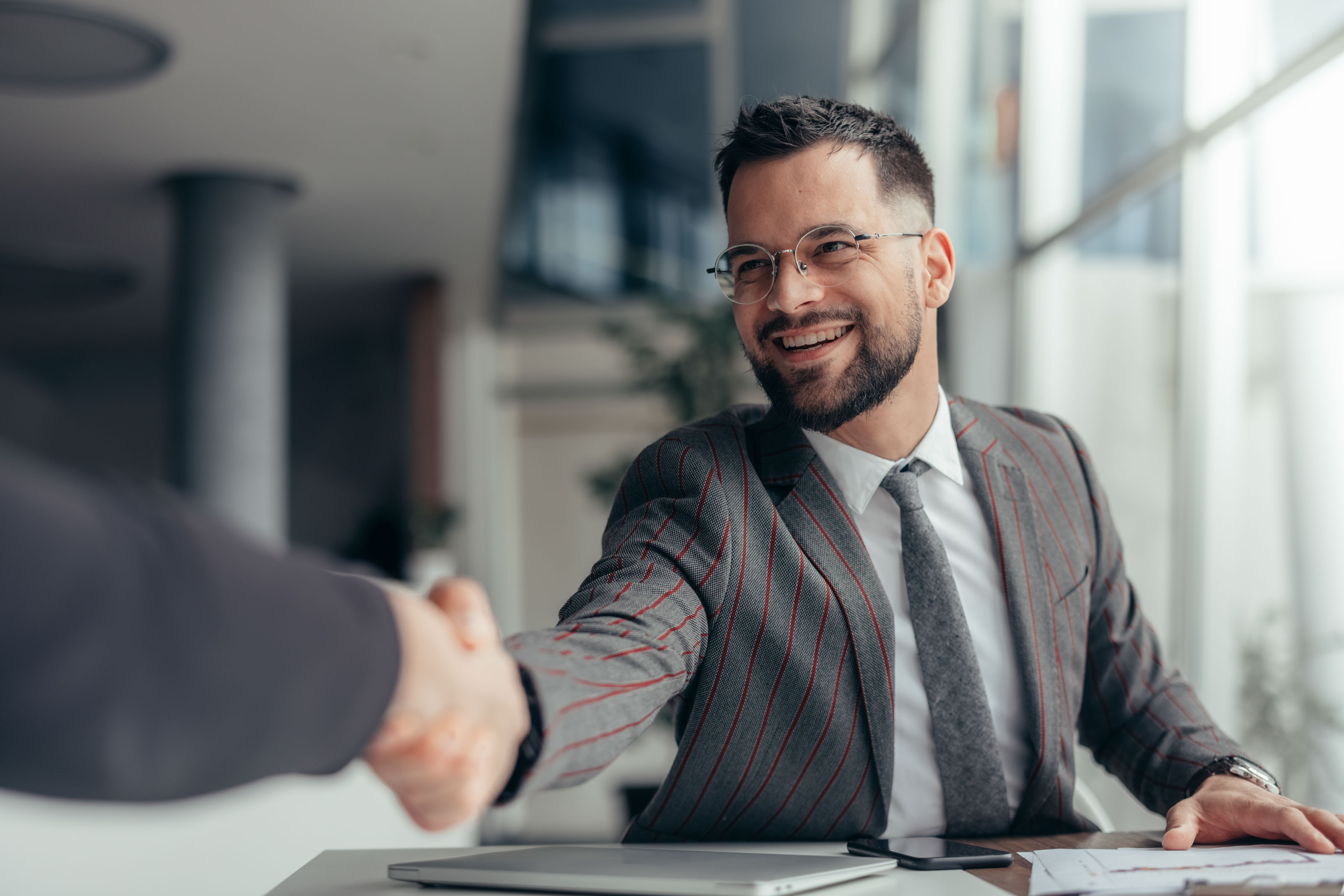 Bearded man in a grey pinstripe suit shaking hands across a desk
