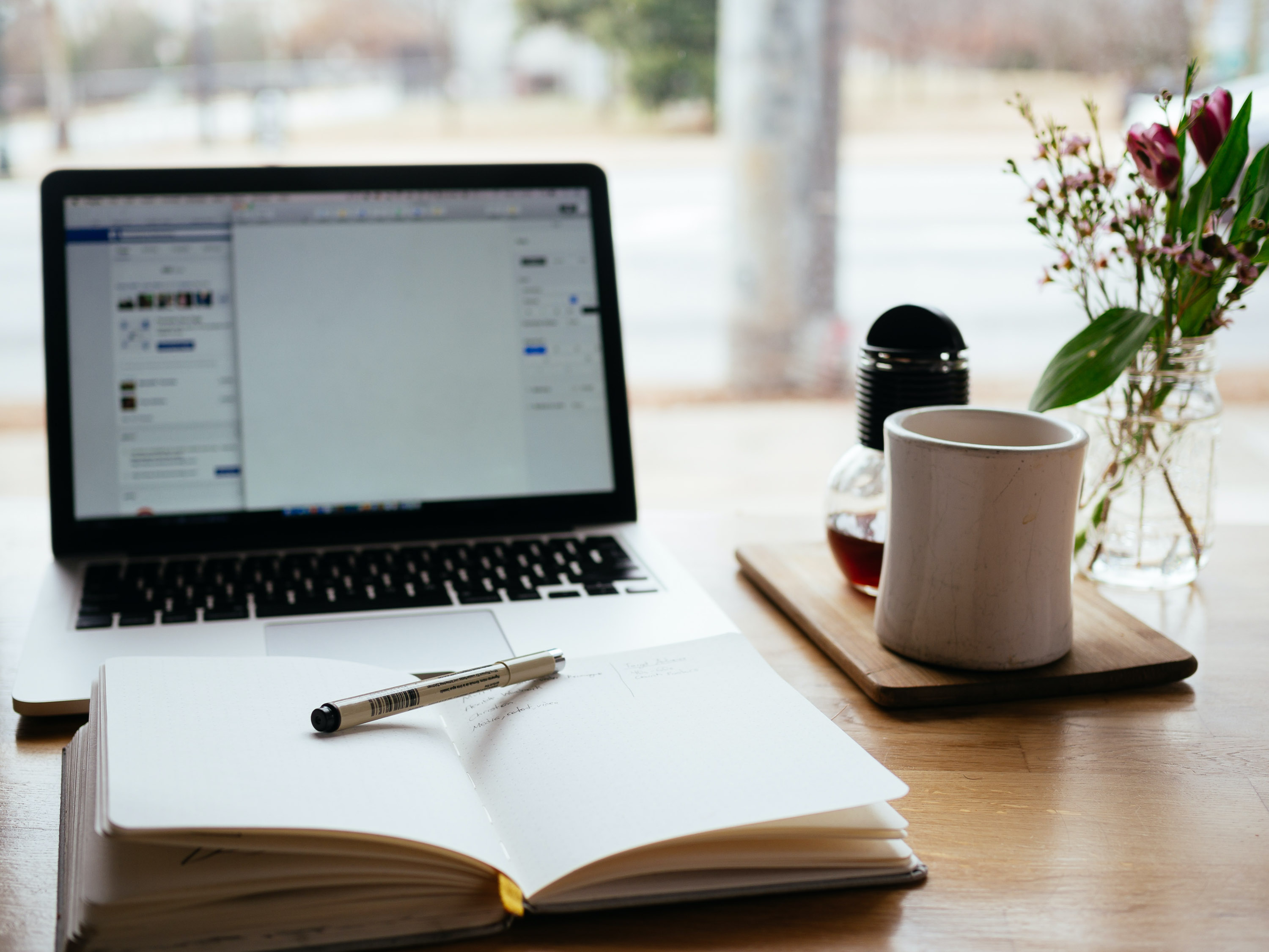 Open laptop and blank notebook on a wooden table by a window