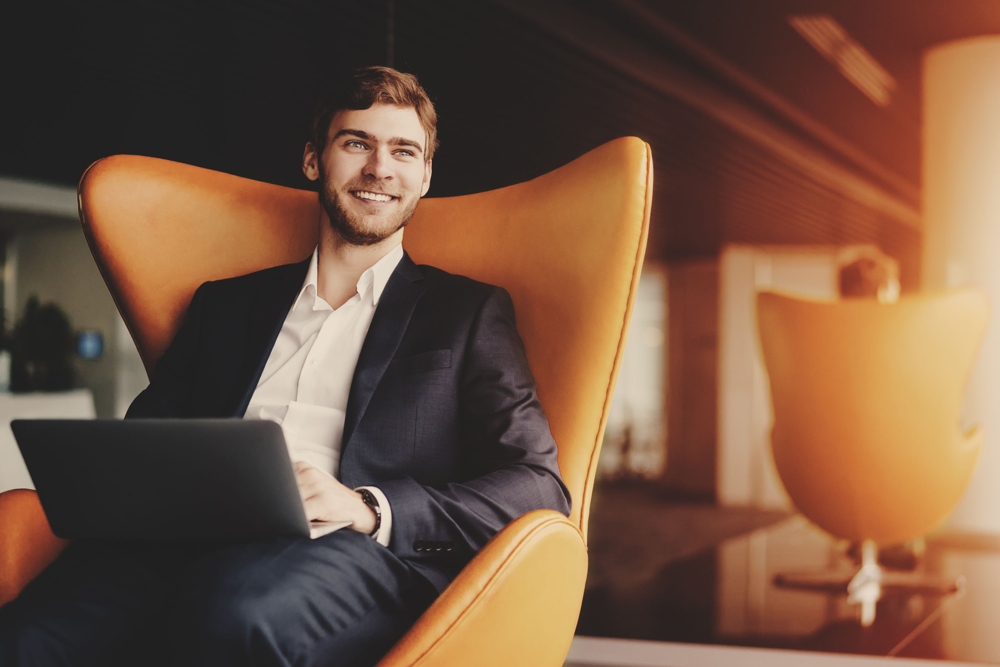 Smiling man in navy suit with a laptop in an orange lounge chair