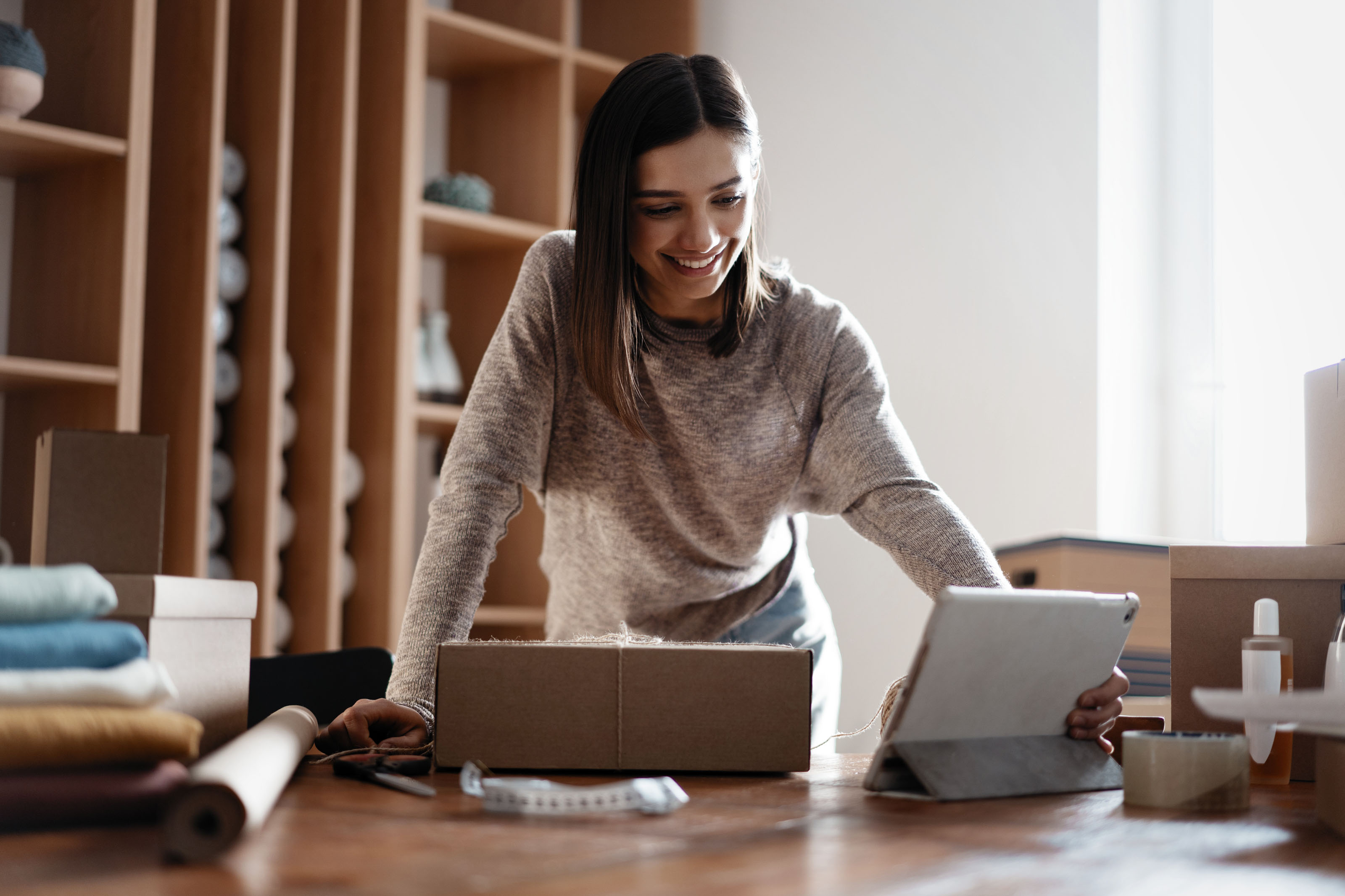 Woman in an oatmeal sweater packing a cardboard box while looking at a tablet