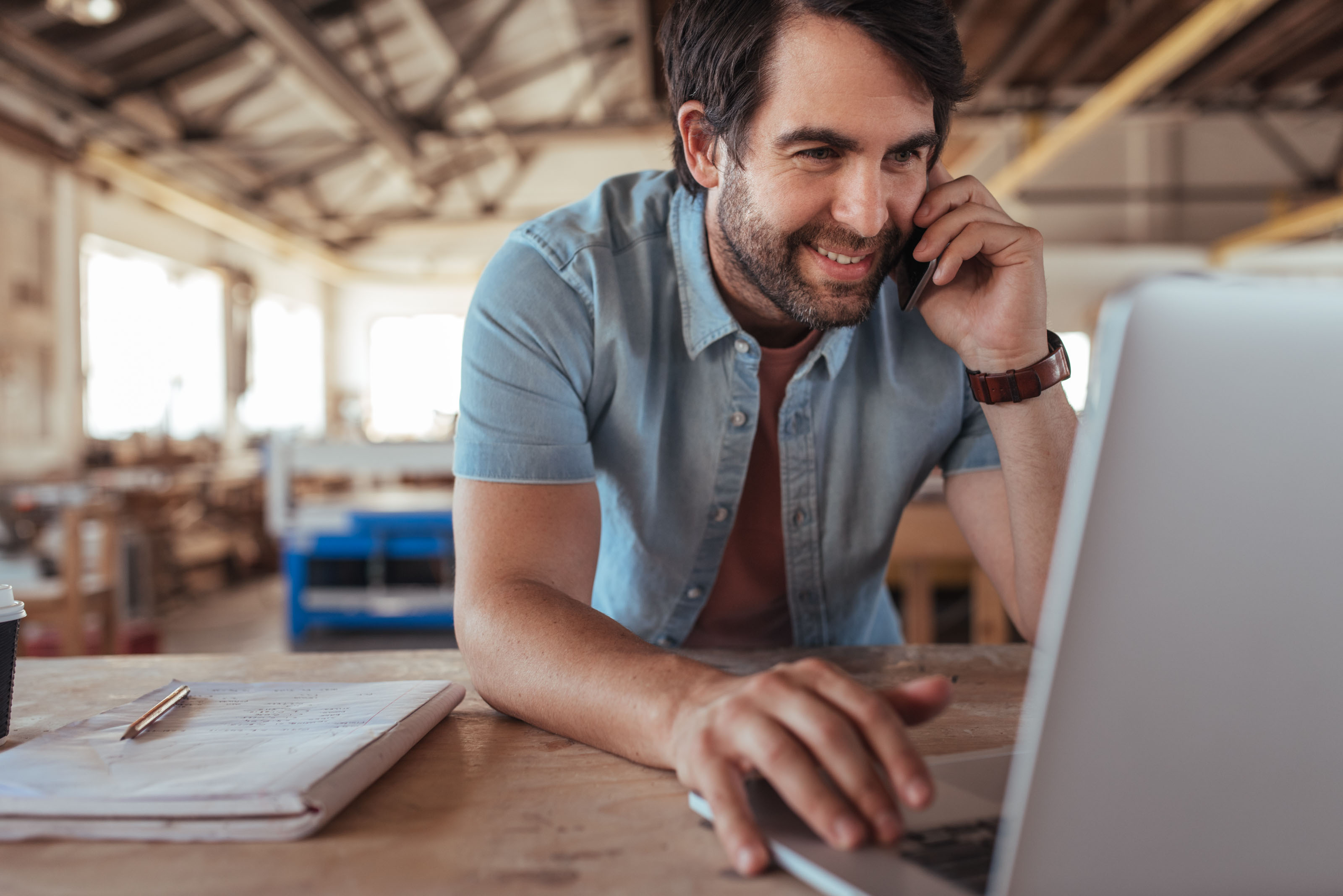 Smiling bearded man on a phone call beside a laptop in a workshop