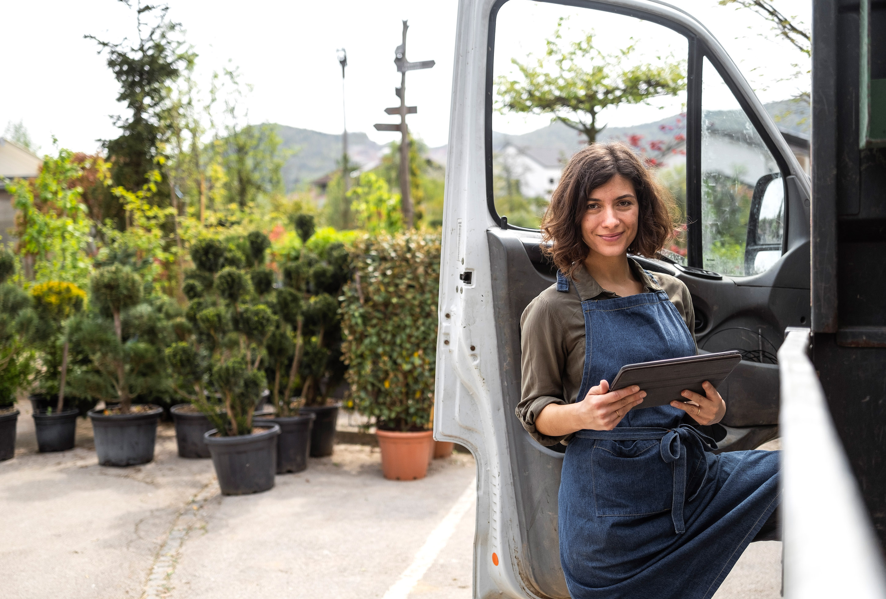 Nursery worker in denim apron holding a tablet beside a van