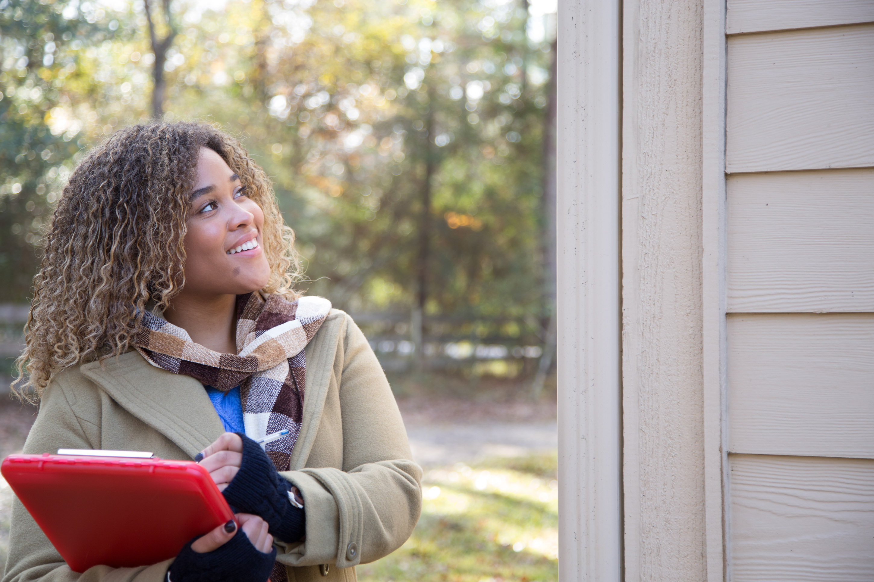A smiling woman in a camel coat holds a red tablet while looking up at the siding of a house during a home service visit