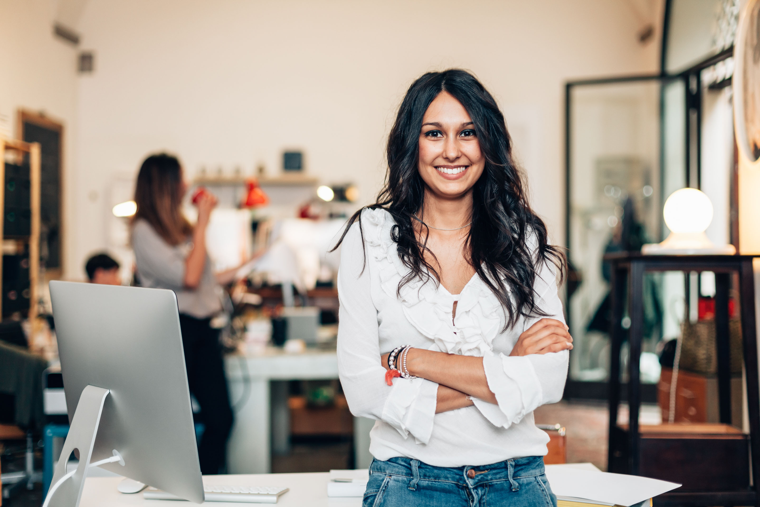 Smiling woman in a white ruffled blouse standing with arms folded at a desk