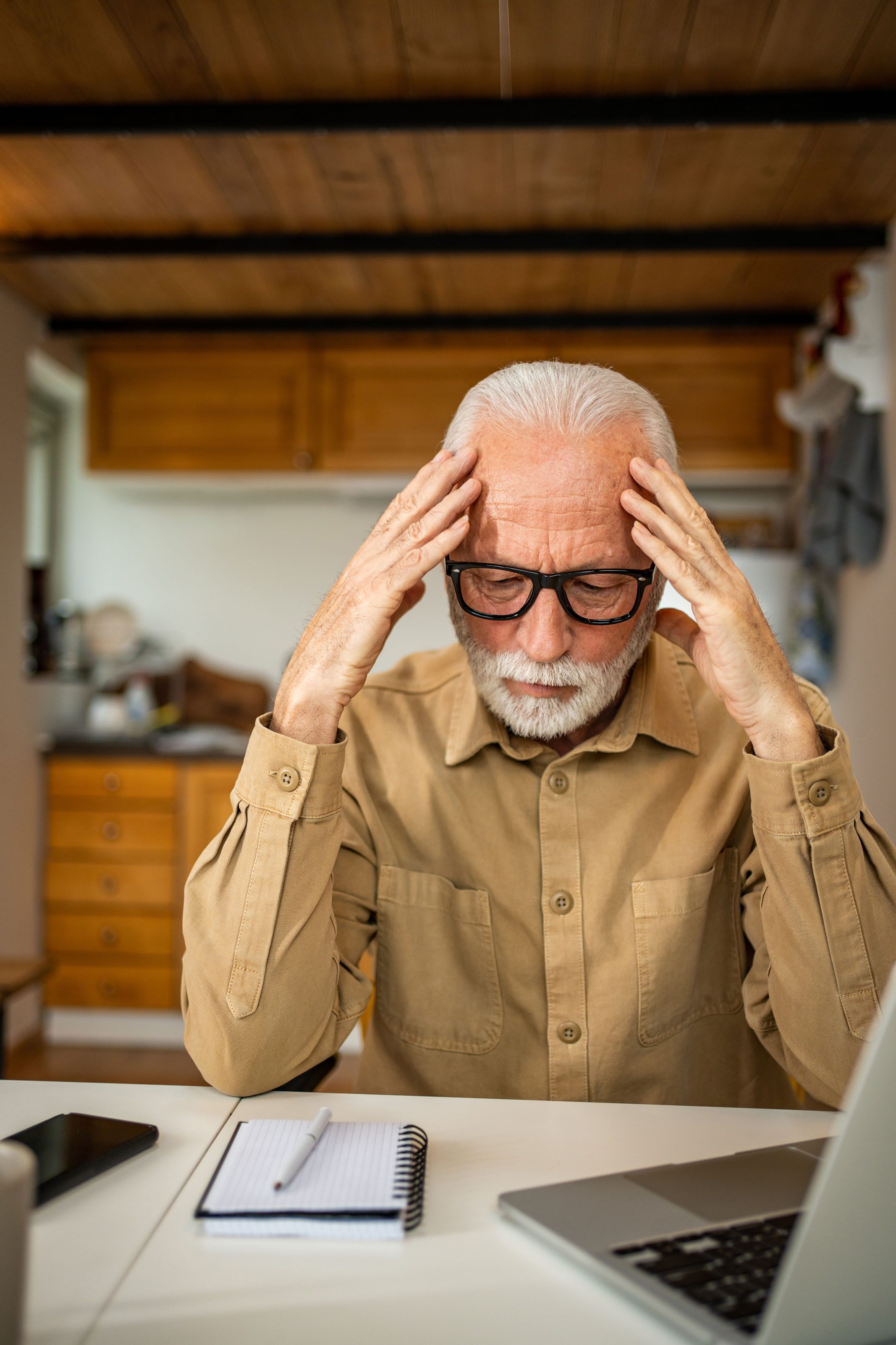 Older bearded man in a tan shirt holding his head at a kitchen table