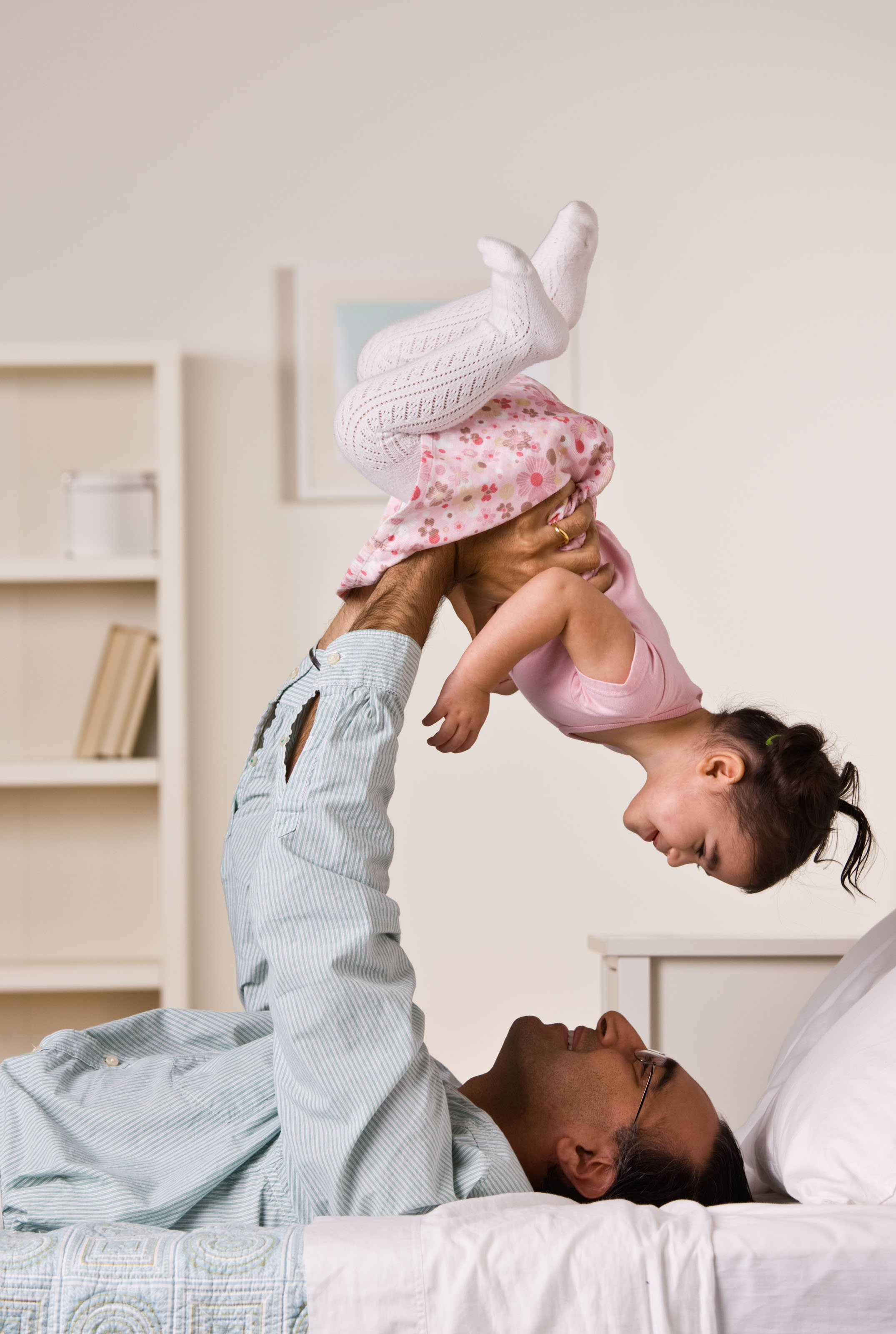 Father lying on a bed lifting a toddler girl overhead