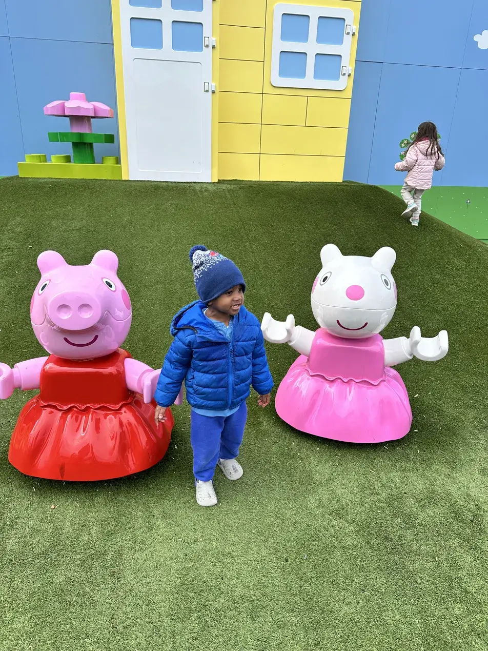 Black toddler in blue jacket walking between Peppa Pig and Suzy Sheep at the LEGOLAND New York Peppa Pig playground