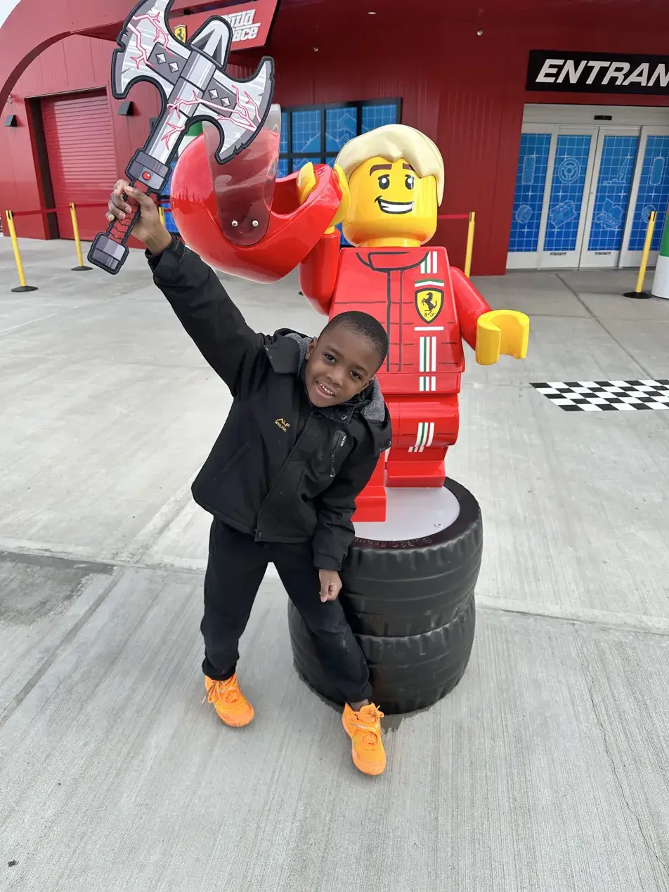 Black boy in orange sneakers posing with a LEGO sword next to the LEGO Ferrari Build and Race statue at LEGOLAND New York