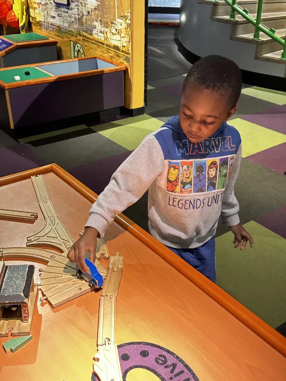 Young Black boy in a Marvel hoodie playing with a wooden train track exhibit at Maryland Science Center Baltimore