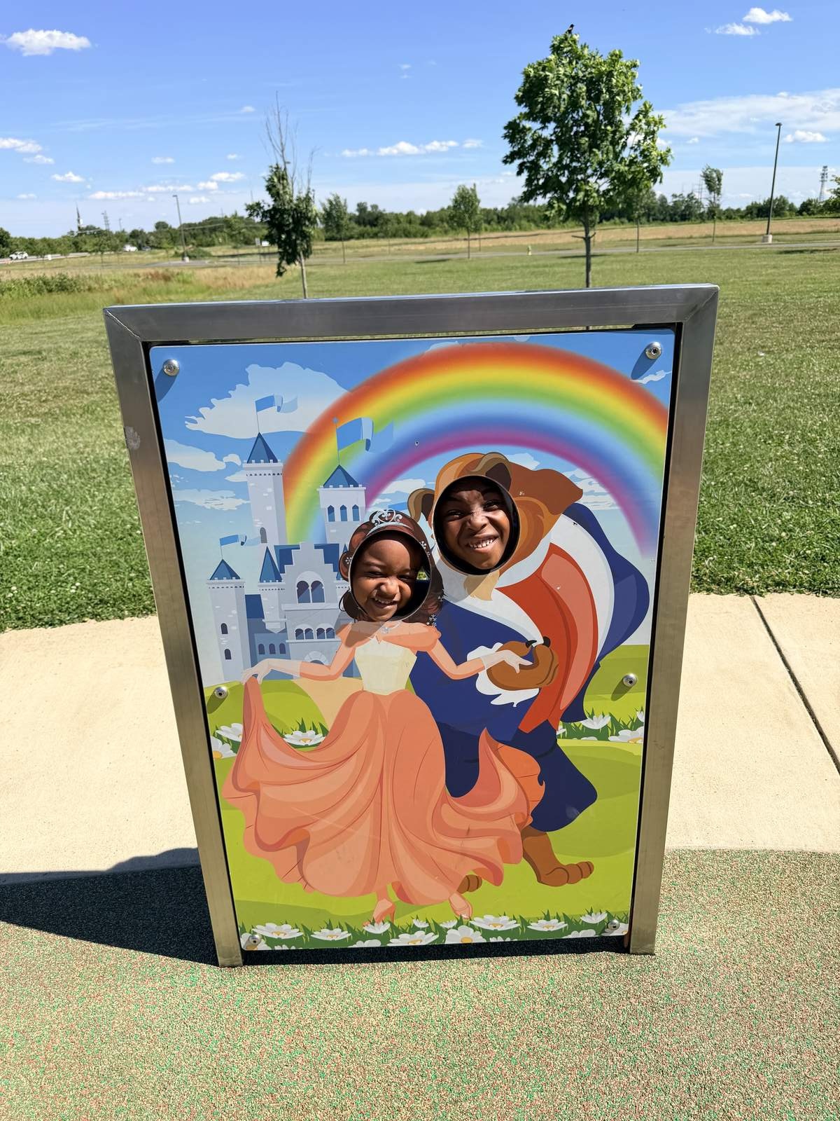 Two children smiling through a princess and prince photo cutout board with a castle and rainbow at Sophie and Madigan's Playground in Frederick, Maryland Two children smiling through a princess and prince photo cutout board with a castle and rainbow at Sophie and Madigan's Playground in Frederick, Maryland