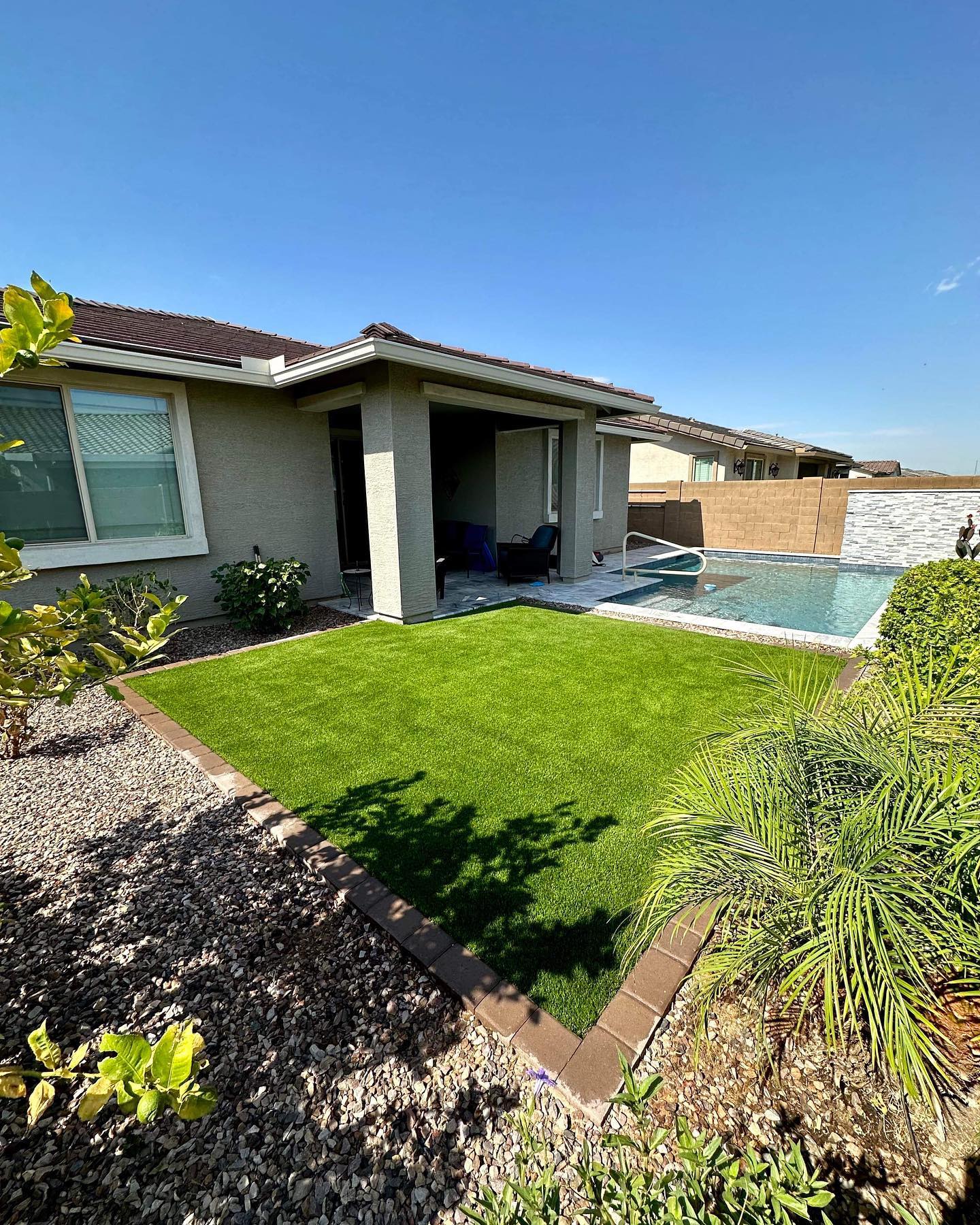 A backyard scene under a clear blue sky, featuring a rectangular artificial grass lawn bordered by brown pavers, surrounded by gravel landscaping, a covered patio of a house, and a swimming pool with a stone feature wall.