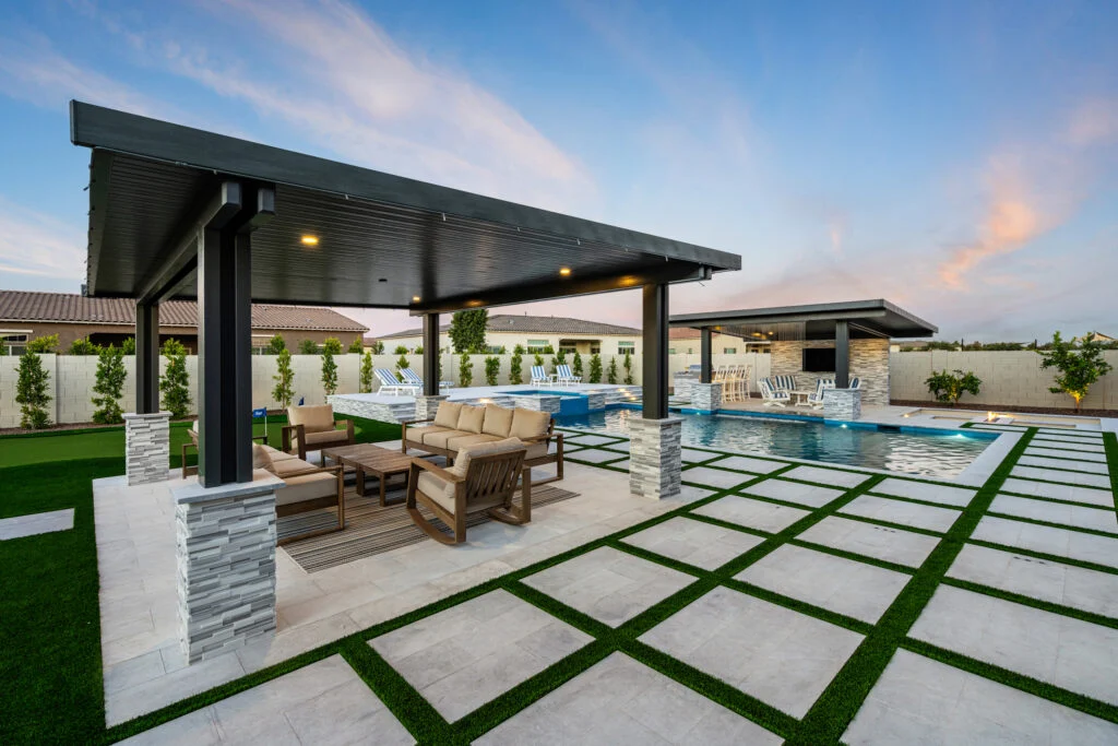 A modern backyard oasis at dusk, featuring a large dark-framed covered patio with stone-accented columns and outdoor seating, stepping stone pavers set in artificial turf, and a swimming pool flanked by an additional covered cabana structure under a twilight sky.