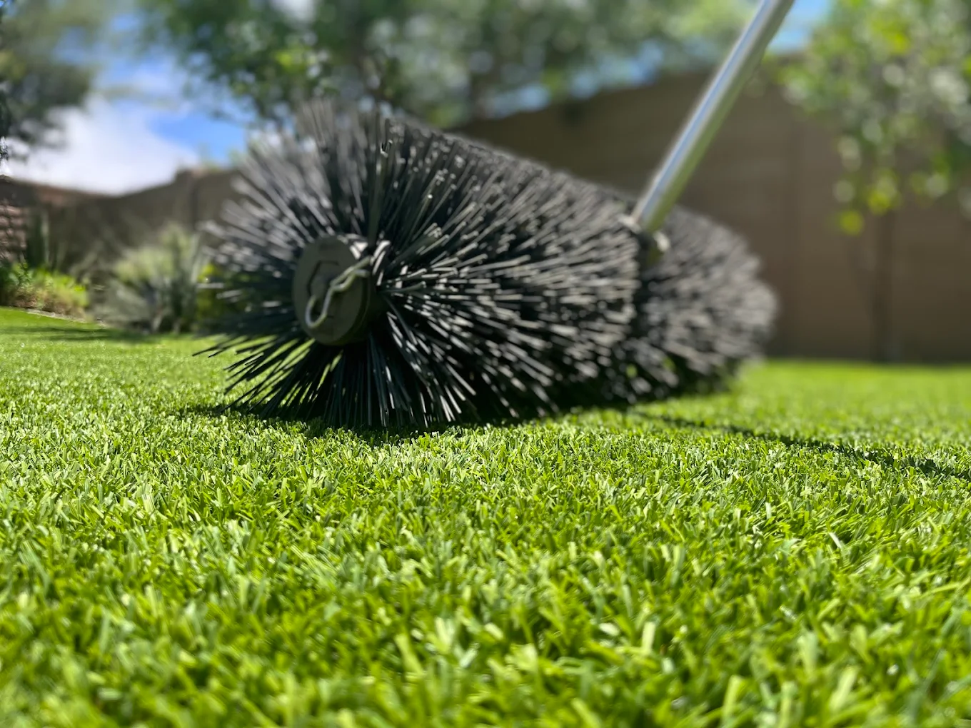 A cylindrical turf broom with stiff black bristles and a metal handle is being rolled across a vibrant green artificial grass lawn outdoors on a sunny day.