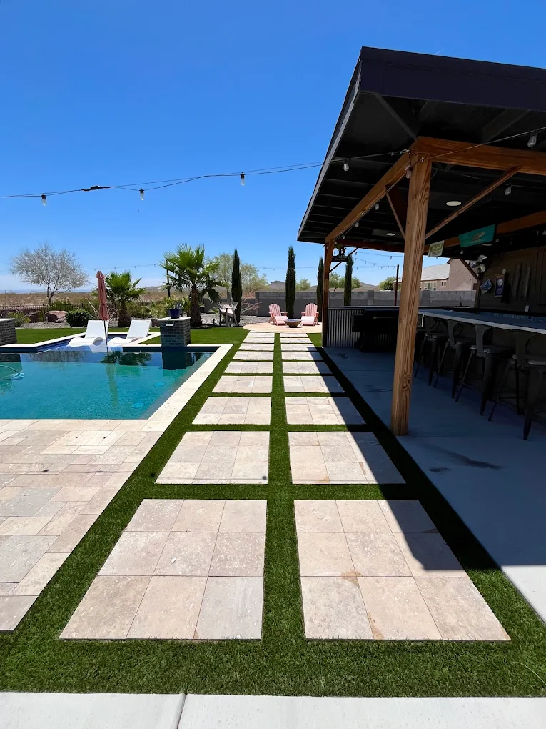 A luxurious outdoor patio featuring a stone fireplace with a fire, comfortable seating, and a swimming pool, next to a manicured garden at sunset.