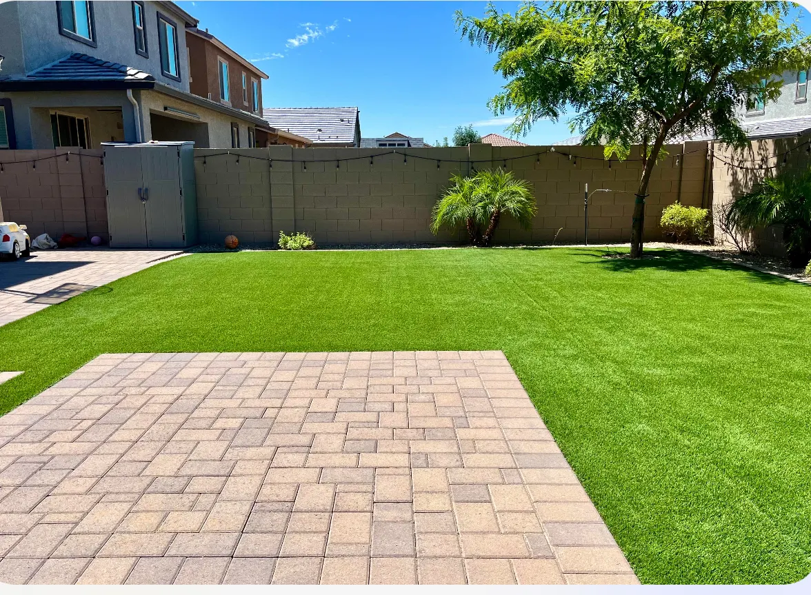 A bright backyard featuring a neatly manicured artificial grass lawn, a square interlocking paver patio in the foreground, a tall block wall fence string-lit with patio lights, and neighboring multi-story homes under a clear blue sky.